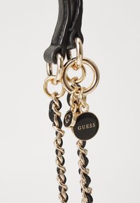 Schwarzer Handtaschenriemen mit ineinander verschlungenen Ketten links und goldenen Beschlägen, Lederakzenten und einem runden Anhänger mit der Marke "GUESS".