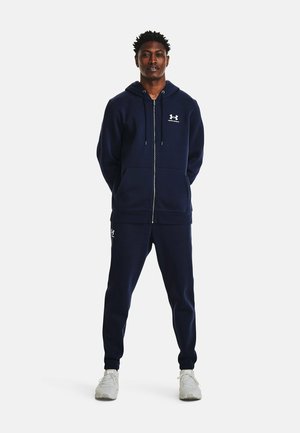 Sudadera con cremallera de color azul marino con bolsillos tipo canguro y pantalones de chándal a juego. La tela parece suave, con un logotipo en el pecho y en el muslo. Acentos en blanco.