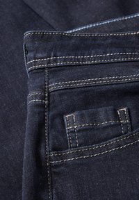 Tissu en denim bleu foncé avec de multiples coutures et poches, accompagné de surpiqûres beige contrastées. Texture lisse avec une légère brillance.