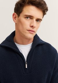 Marineblauer, gerippter Strickpullover mit einem halben Reißverschluss am Kragen. Weiße Innenschicht sichtbar am Halsauschnitt. Weiches Material, taillierte Passform.