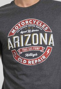 T-shirt gris avec un graphique vintage circulaire, portant "Arizona" en jaune gras et des accents rouges. Le texte inclut "Motos" et "Ancienne Réparation."