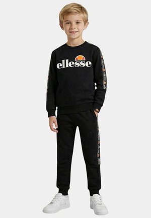 Garçon debout et souriant, portant un sweat-shirt noir Ellessee et un pantalon de survêtement assorti avec des baskets blanches, sur un fond uni.