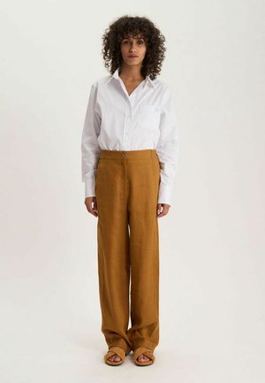 Femme aux cheveux bouclés portant une chemise blanche boutonnée, un pantalon large marron et des sandales beige, debout devant un fond uni.