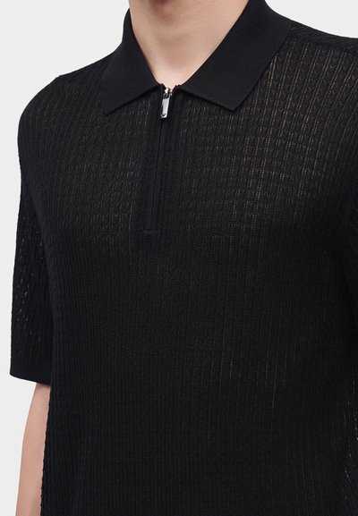 Polo en maille noire à manches courtes avec motif texturé et col zippé sur le devant, porté par une personne cadrée au niveau du cou et de la taille.