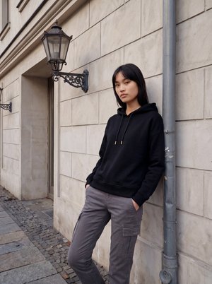 Jeune femme en sweat à capuche noir et pantalon cargo gris, appuyée contre un mur en pierre près d'un lampadaire décoratif dans un environnement urbain.