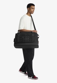 Μαύρη τσάντα duffel από ανθεκτικό ύφασμα, με μπροστινή τσέπη με ιμάντες υφασμάτινους, διπλές χειρολαβές στην κορυφή και αποσπώμενο ιμάντα ώμου.