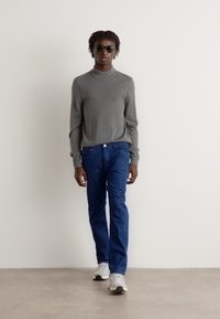 Sivi rolak jumper, plave traperice i bijele tenisice. Model nosi sunčane naočale; stoji protiv jednostavne svijetle pozadine.