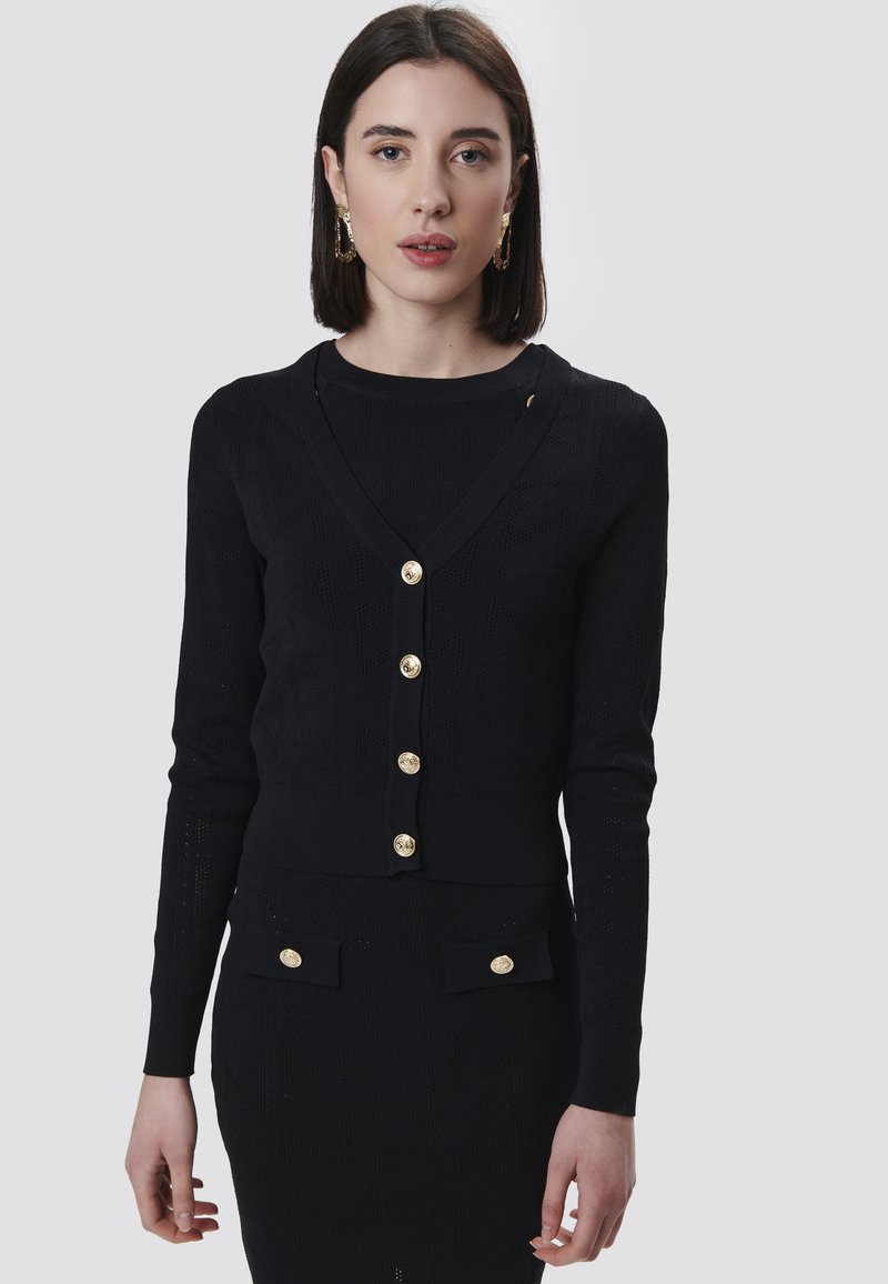 Schwarze Strickjacke mit V-Ausschnitt, langen Ärmeln und goldenen Knöpfen. Hat ein strukturiertes Muster und ein kurzes Design, kombiniert mit einem passenden Rock.