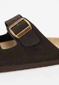 Sandale en daim marron avec une large bride, un design découpé et une boucle en laiton. La semelle intérieure est beige et texturée pour le confort.