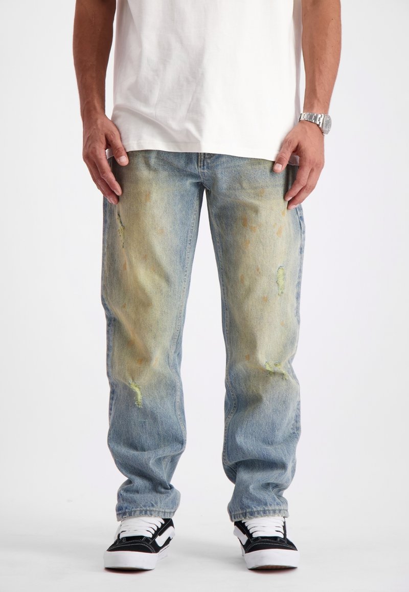 Man in versleten, gescheurde blauw jeans met gele vlekken en zwart-witte sneakers, staande tegen een witte achtergrond.