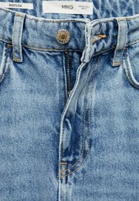 Jean en denim bleu avec fermeture éclair ouverte, bouton en métal, passants de ceinture et étiquettes de marque visibles "MATILDA" et "MNG" à l’intérieur de la ceinture.