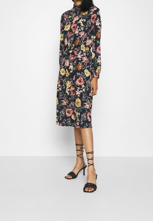 Žena v dlouhých rukávech tmavě modrých šatech s barevným květinovým vzorem a černými sandály na podpatku s pásky, stojící před jednobarevným pozadím.