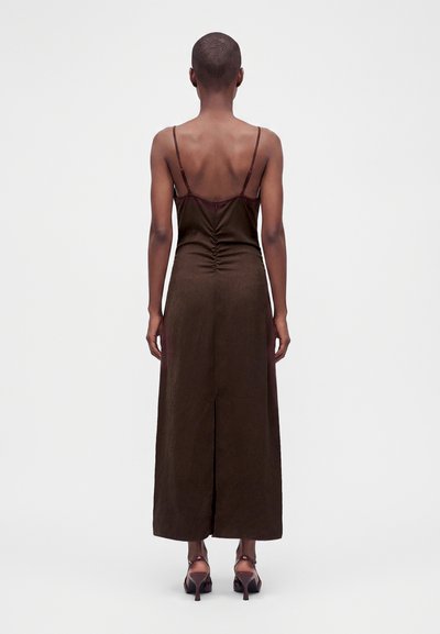 Personne aux cheveux courts portant une longue robe marron foncé sans manches avec de fines bretelles et un dos ouvert, debout, de dos sur un fond uni.