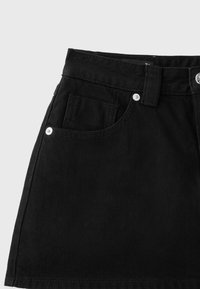 Jupe en denim noire avec poches avant, passants de ceinture et détails de rivets argentés sur fond gris.