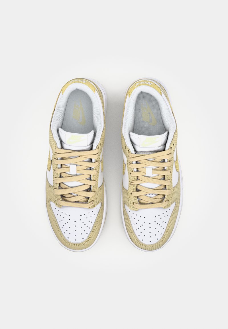 Weiße und beige Nike-Sneaker mit durchbrochener Verzierung, strukturierten Materialien und flachen Schnürsenkeln, versehen mit einem Logo an der Zunge und an der Seite.