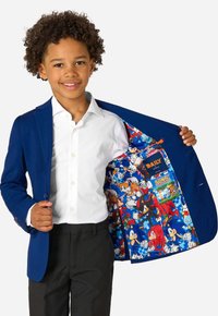 Zils žakets, kas izgatavots no gludas auduma, ar krāsainu komiksu varoņu printa oderi. Uzvilkts virs balta krekla un melniem biksēm.