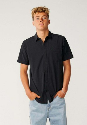 Ung mand med krøllet hår iført en sort kortærmet skjorte med knapper og lyseblå jeans, stående med hænderne i lommerne.