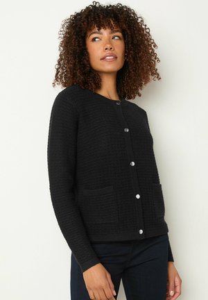Schwarze strukturierte Strickjacke mit rundem Ausschnitt, frontalen Knöpfen und zwei aufgesetzten Taschen. Der Stoff hat ein geripptes Muster.