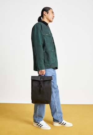 Schwarzer Rolltop-Rucksack mit verstellbarem Verschluss, getragen von einer Person in einer grünen Jacke und blauen Jeans, vor einem gelben Hintergrund.