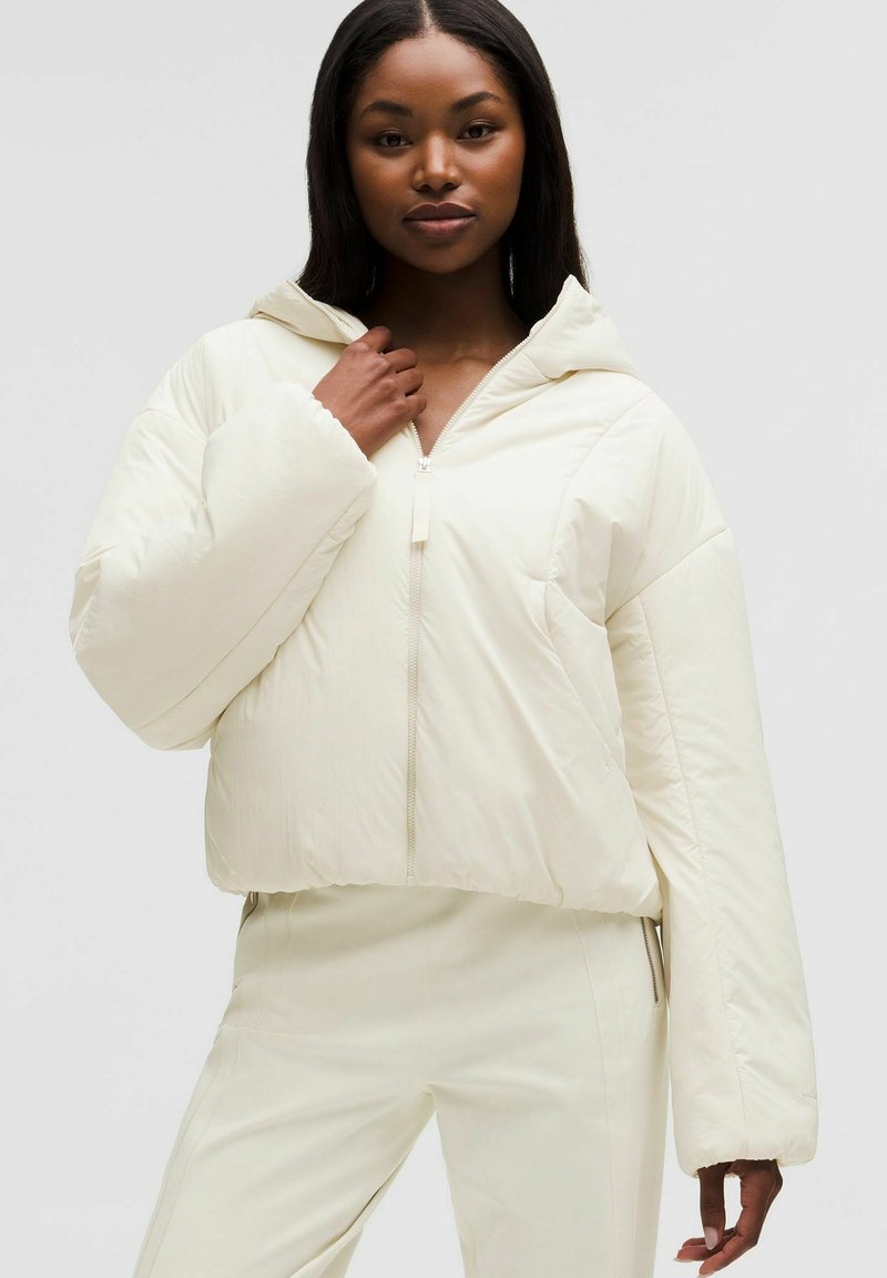 Nainen pukeutuneena kermanväriseen toppatakkiin, jossa on huppu, sekä yhteensopiviin housuihin, poseeraa yhdellä kädellä takin vetoketjun lähellä yksinkertaisen taustan edessä.