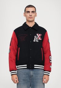 Jachetă varsity roșie și neagră cu închidere cu nasturi, manșete dungi, logo-uri brodate și aplicații pe mâneci. Fabricată dintr-un material cu o finisare netedă.