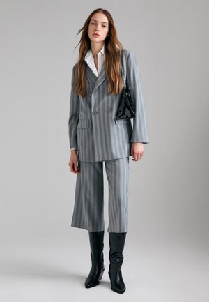 Femme portant un blazer croisé à rayures grises et un pantalon large raccourci assorti, des bottes noires et portant un petit sac noir.