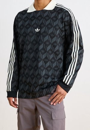 Person iført sort Adidas langærmet skjorte med hvid krave, hvide striber på ærmerne og geometrisk logomønster, kombineret med grå bukser.