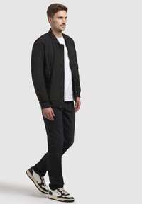 Schwarze Bomberjacke mit Reißverschlusstaschen, getragen über einem weißen T-Shirt, kombiniert mit schwarzen Jeans und schwarz-weißen Sneakern. Glatte Textur.