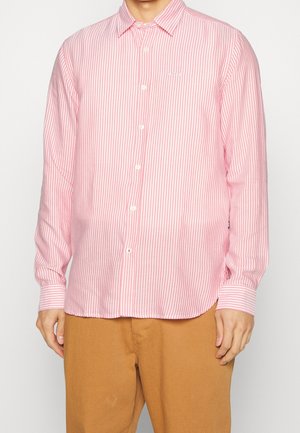 Mężczyzna ubrany w różowo-białą koszulę z pionowymi paskami i długimi rękawami oraz brązowe spodnie.