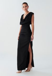 Zwarte avondjurk met een gedrapeerde halslijn, gerimpelde taille en hoge split, gemaakt van glad materiaal; gecombineerd met nude riempjeshakken.