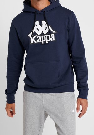 Mężczyzna ubrany w granatową bluzę z kapturem Kappa z białym logo oraz szare spodnie dresowe Champion, stojący z luźno opuszczonymi ramionami na jasnym tle.