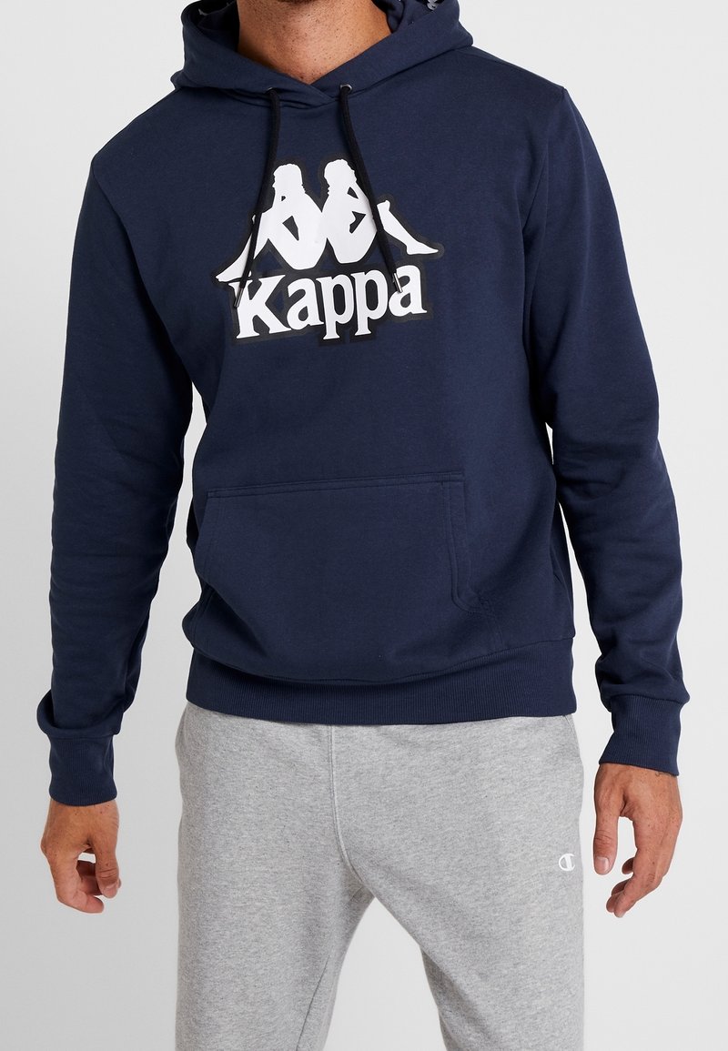 Homme portant un sweat à capuche Kappa bleu marine avec un logo blanc et un pantalon de survêtement gris Champion, debout, les bras relâchés, sur un fond clair.