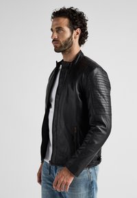 Zwarte leren jas met getextureerde schouderaccenten, voorrits, staande kraag en slim fit. Gecombineerd met een wit shirt en blauwe jeans.