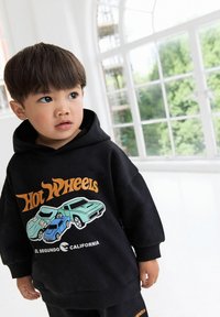 Young child wearing a black hoodie with Hot Wheels cars and text "El Segundo California," standing by a large window with greenery outside.