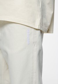T-shirt oversize color beige chiaro abbinato a pantaloni coordinati, con un ricamo sottile della scritta "PEGALUR" in una leggera tonalità di lavanda. Tessuto in cotone morbido.