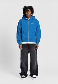 Blauwe rits-hoodie van een katoenmix, met voorzakken en geribde zoom. Gecombineerd met losse zwarte jeans en zwarte sneakers.