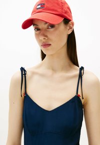 Gorra roja con el logo bordado "TOMMY JEANS", usada con un vestido azul marino que presenta lazos ajustables y un delicado borde festoneado.