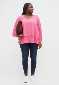 Γυναίκα με ροζ μπλούζα με στρώσεις, σκούρο τζιν, λευκά αθλητικά παπούτσια, κρατώντας καφέ τσάντα, κοιτώντας προς τα πλάγια σε απλό λευκό φόντο.