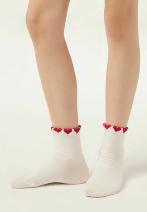 Caviglie con calzini bianchi decorati con motivi a forma di cuore rossi lungo i bordi superiori, in piedi su una superficie chiara.
