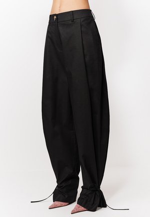 Czarne, szerokie spodnie z wysokim stanem i wiązaniami przy mankietach, noszone z różowymi butami na spiczastym nosku, na prostym białym tle.