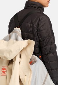 Musta puffatakki korkean kauluksen kanssa, kerrostettuna vaalean kerman ja harmaan ulkohaalarin kanssa. Ominaisuuksiin kuuluu säätöremmit ja näkyvät logot.