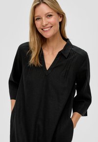Glunderende blonde vrouw in een zwarte V-hals jurk met driekwartmouwen, met haar handen in de zakken, staand tegen een effen lichte achtergrond.