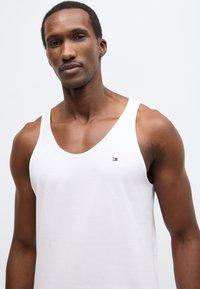 Maiou alb din bumbac cu decolteu rotund, având un mic logo multicolor pe pieptul stâng și o textură netedă, ușoară.