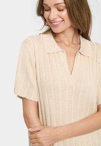 Beige bordázott kötött polo ruha rövid ujjakkal, V-nyakkal és diszkrét függőleges csíkokkal, puha textúrával és laza fazonnal.