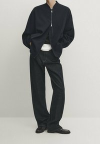 Henkilö, joka pukeutuu mustaan vetoketjulliseen pommikaukovaatteeseen, tummiin leveisiin farkkuihin, valkoisen paidan alla ja mustiin nahkakenkiin, seisoo kädet taskuissa.