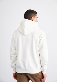 Sudadera blanca con una capucha amplia, tejido texturizado, corte relajado, puños y cinturilla de canalé, combinada con pantalones marrones.