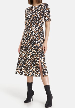 Femme portant une robe longueur genou avec un motif abstrait noir, blanc et marron et une fente latérale, associée à des bottines noires à talons hauts.