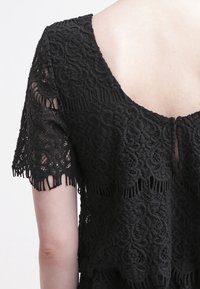 Svart spetstopp med korta ärmar, invecklade blommönster, en vid halsringning och fransdetaljer i nederkant och ärmslut.