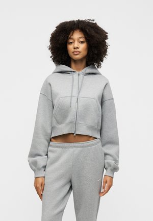 Kvinde med krøllet hår iført grå cropped hættetrøje og matchende joggingbukser mod enkel lys baggrund.