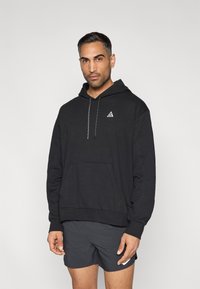 Μαύρο φούτερ με κουκούλα και μπροστινή τσέπη, με μικρό τριγωνικό λογότυπο και υφασμένα κορδόνια. Φορεμένο με σκούρα σορτς.